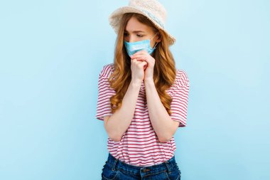 Yazlık şapkalı dua eden bir kız yüzüne tıbbi koruyucu bir maske takıyor, dua ediyor, ellerini kavuşturuyor, mavi bir arka planda. Karantina, koronavirüs, yaz tatili konsepti
