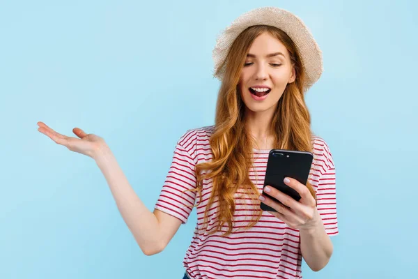 戴着夏帽的美丽的快乐姑娘 用手机 站在孤零零的蓝色背景上 — 图库照片