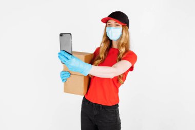 Tıbbi lastik eldiven ve tıbbi maske takan üniformalı bir kurye karton kutular tutuyor ve beyaz arka planda bir cep telefonu kullanıyor. Çevrimiçi mağazalar, koronavirüs, karantina
