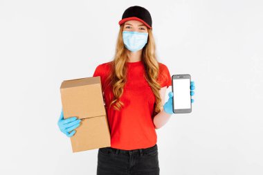 Plastik eldiven ve tıbbi maske takan üniformalı bir kurye, karton kutular tutuyor ve beyaz arka plandaki boşluğun bir kopyası için boş bir cep telefonu ekranı gösteriyor..