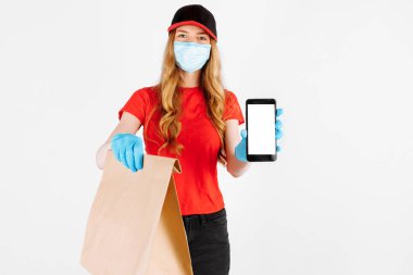 Üniformalı bir kurye, lastik eldiven ve tıbbi maske takıyor, elinde bir torba yiyecek tutuyor ve beyaz arka plandaki boşluğun bir kopyası için boş bir cep telefonu ekranı gösteriyor..