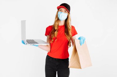 Plastik eldiven ve tıbbi maske takan üniformalı bir kurye, elinde beyaz arka planda yiyecek dolu bir laptop ve kese kağıdı tutuyor. Çevrimiçi dağıtım