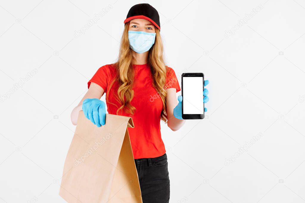 A uniformed courier, wearing rubber gloves and a medical mask, holds a paper bag of food and shows a blank mobile phone screen for a copy of the space on a white background.