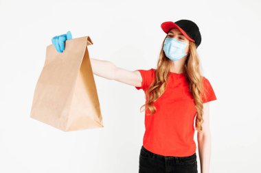Kırmızı şapkalı ve tişörtlü bir kurye, yüzünde tıbbi maske ve beyaz arka planda yiyecek dolu bir kese kağıdı tutan eldivenler. Ev teslimatı, karantina, koronavirüs...