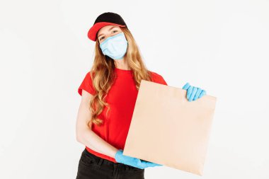 Kırmızı şapkalı ve tişörtlü bir kurye, yüzünde tıbbi maske ve beyaz arka planda yiyecek dolu bir kese kağıdı tutan eldivenler. Ev teslimatı, karantina, koronavirüs...