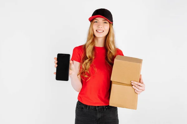 穿着制服的年轻姑娘 手里拿着纸板箱 在白色背景上显示一个空白的手机屏幕 以获取空格的副本 网上购物 — 图库照片