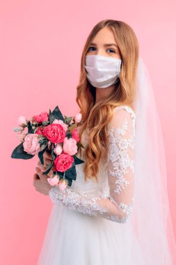 Gelinlik giymiş güzel bir gelin ve yüzünde tıbbi koruyucu bir maske, izole pembe bir arka planda. Düğün, karantina, koronavirüs.