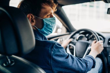 Koruyucu tıbbi maskeli genç bir adam bir arabanın direksiyonunda oturuyor ve şoför kullanıyor. Karantina, koronavirüs