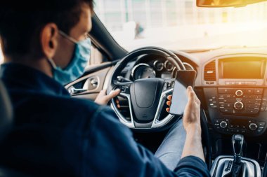 Koruyucu tıbbi maskeli bir adam, direksiyon başında oturuyor, araba sürerken cep telefonu kullanıyor. Karantina, koronavirüs