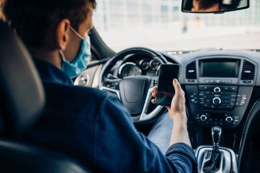 Koruyucu tıbbi maskeli bir adam, direksiyon başında oturuyor, araba sürerken cep telefonu kullanıyor. Karantina, koronavirüs