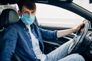 Koruyucu tıbbi maskeli genç bir adam bir arabanın direksiyonunda oturuyor ve şoför kullanıyor. Karantina, koronavirüs