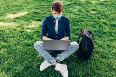 Koruyucu tıbbi maske takan genç bir öğrenci bir dizüstü bilgisayarla parkta çimlerin üzerinde oturuyor. Karantina, koronavirüs
