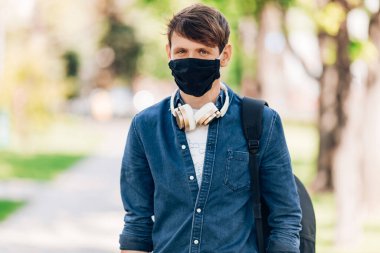 Yüzünde koruyucu tıbbi maske olan genç bir adam boynunda kablosuz kulaklıkla parkta yürüyor. Karantina, koronavirüs