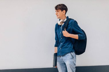 Genç bir adam elinde bir çanta ve dizüstü bilgisayarla beyaz bir duvara karşı duruyor.