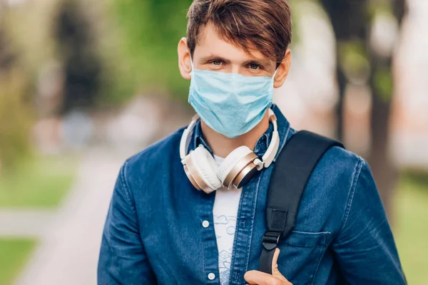 A young man in a protective medical mask on his face walks through the Park with wireless headphones around his neck. Quarantine, coronavirus