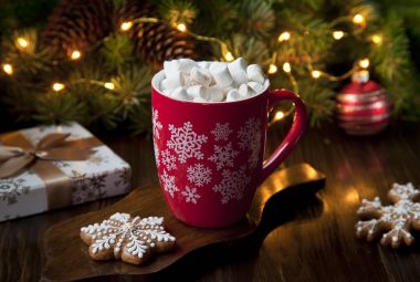 Close-up of a mug with a hot drink, marshmallow and gingerbread. Christmas decor with a bright garland and fir tree. Selective focus, shallow depth of field, background