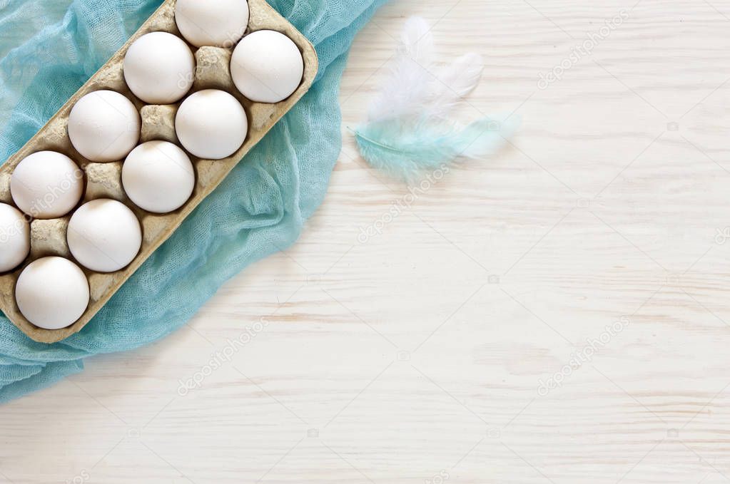 Close-up of white Easter eggs in paper box with blue scarf on wooden table background and feathers