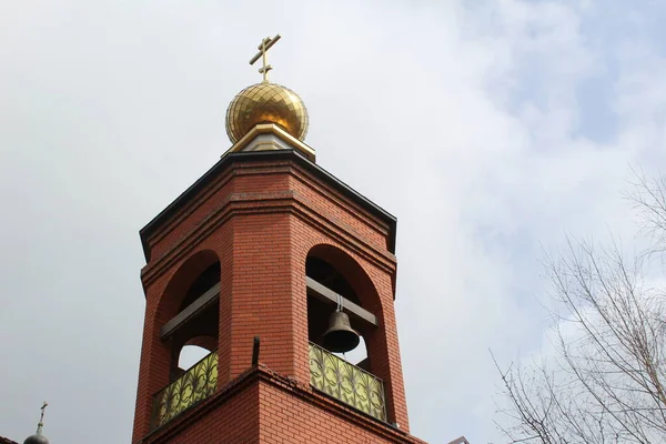 Church bell tower with a Golden dome and a cross against the sky in Russia / photo of the Church in Russia.A bell hangs on the belfry. There is a beautiful dome at the top.It has a large gold cross on it.