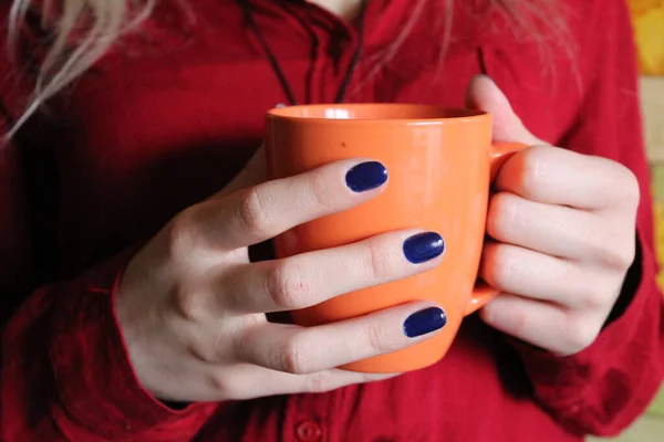 a woman with a Cup of hot tea, coffee in her hands / photo orange Cup,large. it contains tea and coffee. it is held by a woman in a maroon shirt. hands are beautiful. long fingers. the nails are manicured, blue Polish.