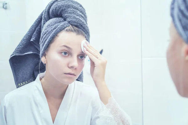 La chica en el baño delante del espejo con una toalla en la cabeza se aplica crema en la cara. enfoque selectivo. grano de película — Foto de Stock