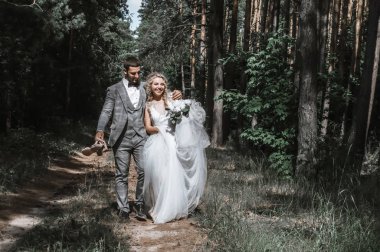 Gelin ve damat ormandan kucaklaşarak geçiyorlar. Yalınayak gelin, damat ayakkabı taşıyor. Güneşli yaz günü.