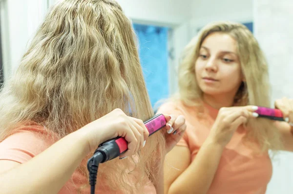 The girl at the mirror straightens curly hair with a hair straightener at home. Selective focus, film grain. — Stock Photo, Image