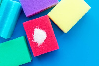 Mavi arka planda çok renkli süngerlerin yanında kırmızı bir bulaşık süngerinin üzerindeki temizlik tozu. Ev ödevi konsepti.