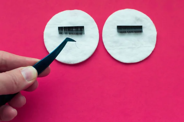 the hand of the eyelash extension master uses tweezers to take an artificial eyelash from a cotton pad on a pink background.