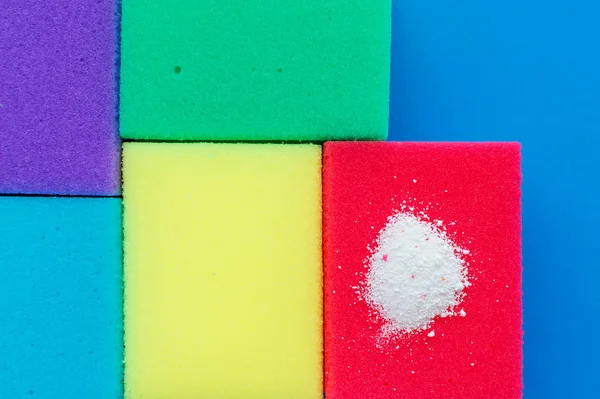 Cleaning powder on a red dishwashing sponge next to multi-colored sponges on a blue background. The concept of homework.