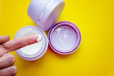 Yaşlı bir adamın elindeki krem, arka plandaki gri yüz kremi kavanozlarına karşı sarı arka planda. Cilt bakımı.