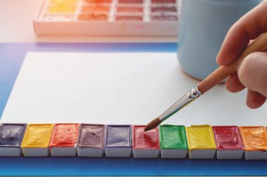 Sanatçının eli beyaz bir kağıt üzerinde bir fırça tutuyor. Suluboya resimler, suyla bardaklar. Yakın plan..