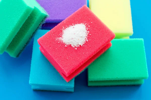 Pó de limpeza em uma esponja de lavar louça vermelha ao lado de esponjas multicoloridas em um fundo azul. O conceito de lição de casa . — Fotografia de Stock
