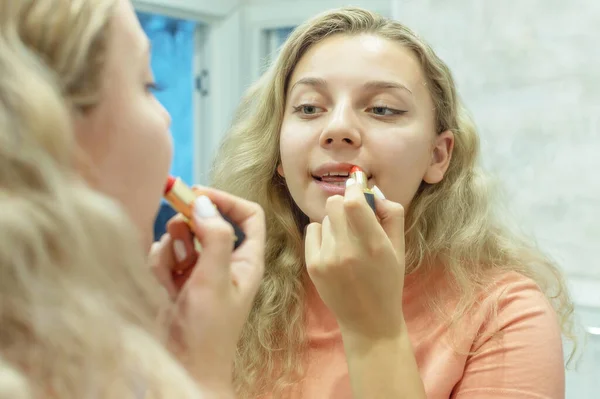 Una chica con el pelo rizado se pinta los labios con lápiz labial rojo delante del espejo en casa. De cerca. Enfoque selectivo, grano de película . — Foto de Stock