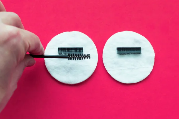 the hand of the eyelash extension master combs artificial eyelashes with a brush on a cotton swab on a pink background.