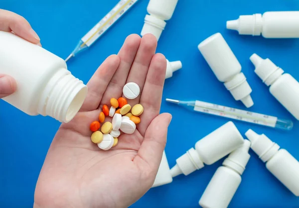 The hand pours a handful of pills into the other hand against a background of nose drops and thermometers on a blue background. Concept of health protection. — 스톡 사진