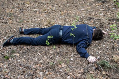 Ormanda cinayet. Mavi gömlekli ve pantolonlu bir adamın cesedi ormandaki ağaçların arasında yerde yatıyor. Bir saldırı kurbanı. Yatay fotoğraf.