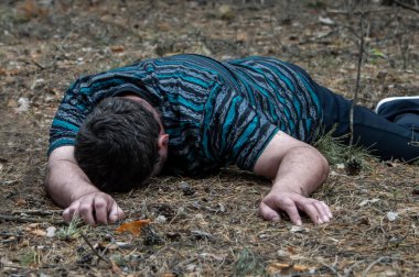 Ormanda cinayet. Mavi tişörtlü ve pantolonlu bir adamın cesedi ormandaki ağaçların arasında yerde yatıyor. Bir saldırı kurbanı. Yatay fotoğraf.