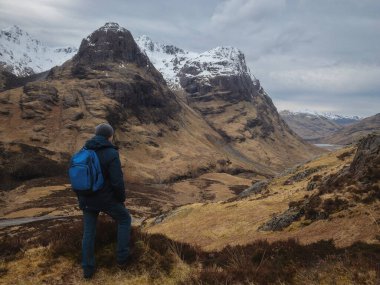 Sırt çantalı bir adam dağdan dağ vadisine bakıyor. Aktif rahatlama, motivasyon ve hedefe ulaşma kavramı. Glencoe, İskoçya, İngiltere.