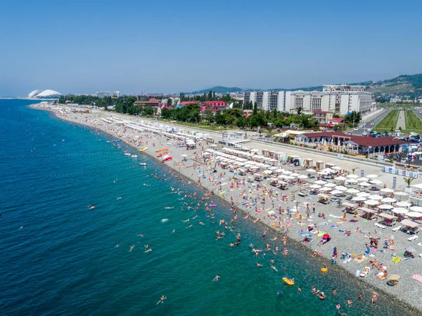 Sochi Olimpiyat Parkı yakınlarındaki plaj, hava manzaralı. İnsanlar güneşleniyor, yüzüyor. Plaj şemsiyeleri, beyaz güneş panjurları. Güneşli yaz günü.