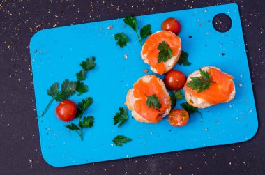 Taze bruschetta somon üstte maydanoz ile. Maydanoz ve domates kiraz sağ tarafında üç kanepeler arasında. Tüm mavi kesme tahtası üzerinde koyu arka plan. Çevresinde dökülen baharat