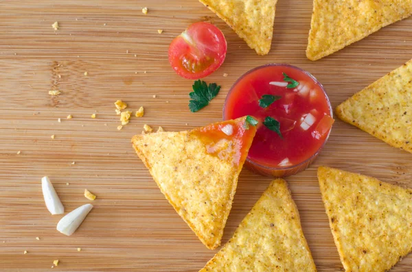 Nachos snack with spicy tamato garlic dip on light brown cutting board and scattered around crumbs, garlic and half of cherry tomato