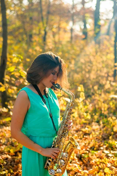 a girl with a beautiful figure and black hair looks away, standing in a yellow autumn park in a long blue dress with an alto saxophone in her hands