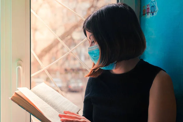 一个黑头发 身穿黑色衣服的女孩 戴着外科口罩坐在医院窗前看书 — 图库照片