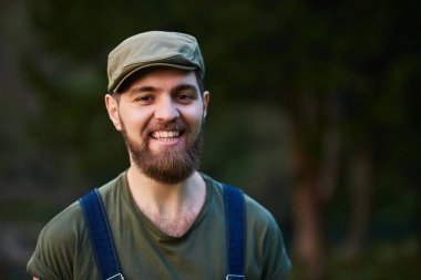 Portrait of handsome smiling bearded man wearing overall standing in summer park  clipart