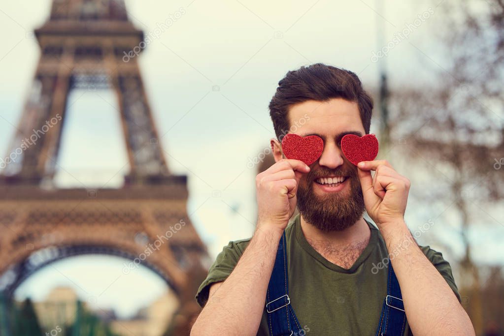 Portrait of handsome smiling bearded man holding red hearts in hands over blurred Eiffel Tower background