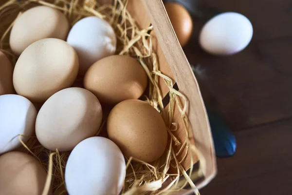 Fresh chicken eggs in wooden box with hay and feathers on wooden background.