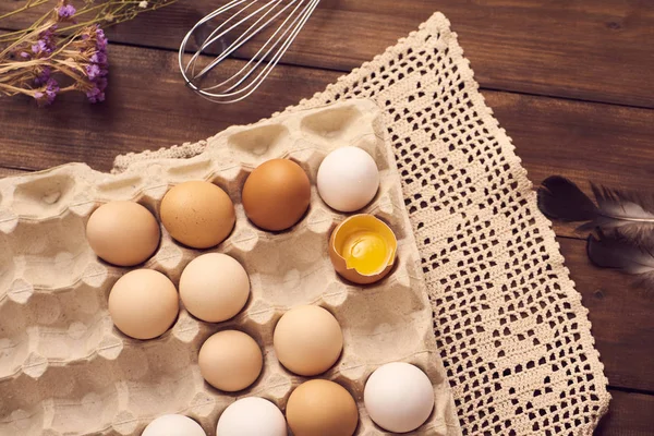 Closeup of broken egg among eggs in carton box with napkin, whisk and flowers on wooden table.