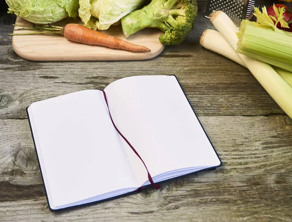 Top view of open recipe book with vegetables on wooden background