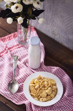 Hububat beyaz kase, kaşık, şişe süt, mutfak havlusu ve koyu kahverengi ahşap arka plan üzerinde cam vazoda güzel çiçekler.