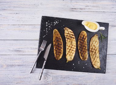 Çatal, bıçak ve sos beyaz rustik arka plan üzerinde siyah arduvaz plaka üzerinde sırlı ızgara patlıcan. Üstten Görünüm